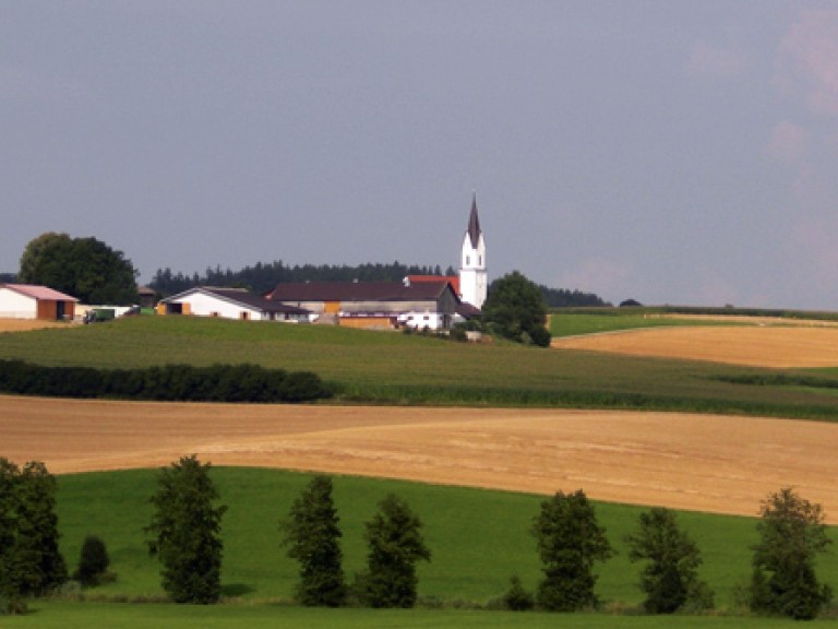 Im Vordergrund ist ein Getreidefeld, im Hintergrund ein Dorf zu sehen.