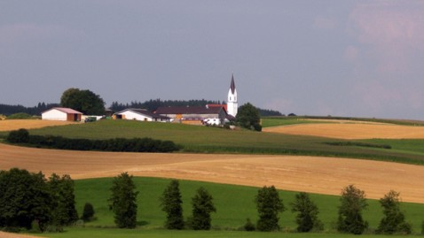 Im Vordergrund ist ein Getreidefeld, im Hintergrund ein Dorf zu sehen.