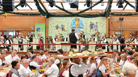 Blick ins voll besetzte Festzelt. Im Hintergrund spielen auf der Bühne die Münchner Oktoberfest Musikanten. Im vordergrund trägt ein Ober ein volles Tablett vorbei.