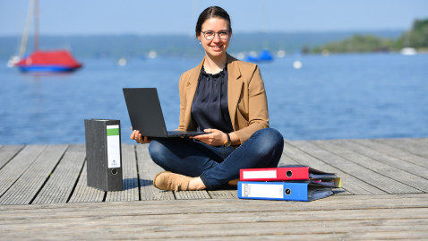 Frau sitzt am See mit Laptop und Ordnern